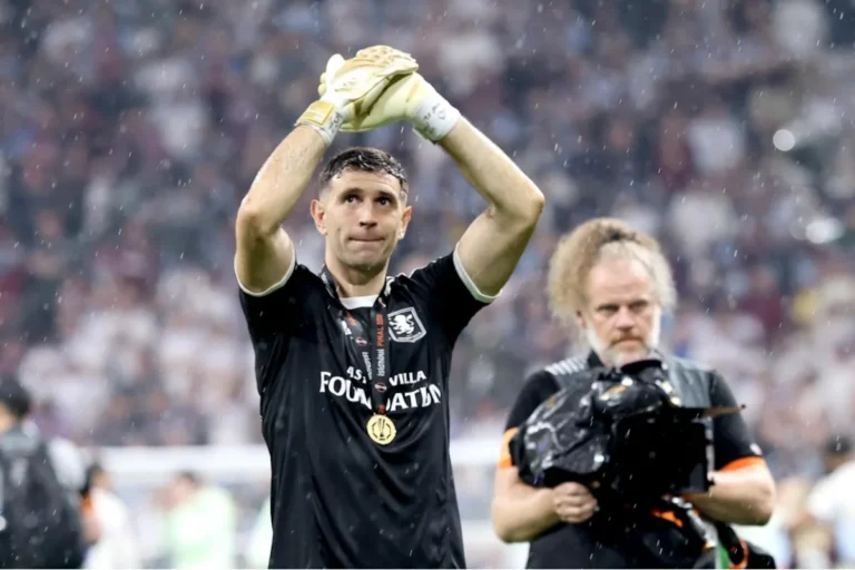 Emiliano Martinez, portiere dell'Aston Villa, alza le mani al cielo sotto la pioggia indossando la maglia nera con medaglia al collo