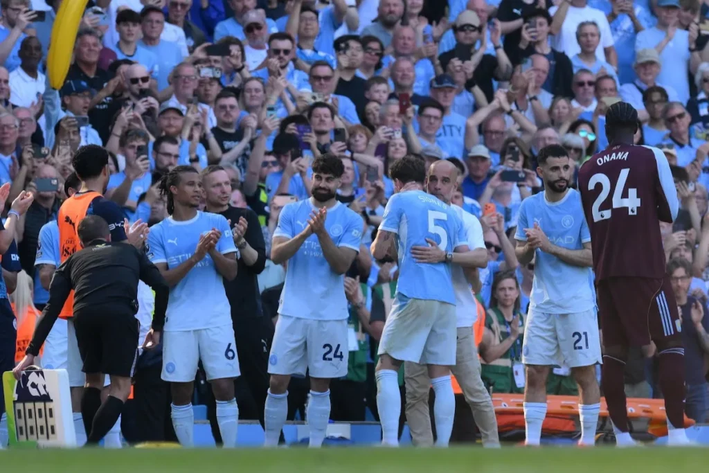 Giocatori del Manchester City in maglia azzurra applaudono durante una partita, con la folla sullo sfondo dello stadio