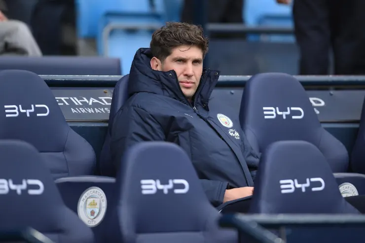 John Stones in panchina con la giacca del Manchester City, seduto in tribuna durante una partita