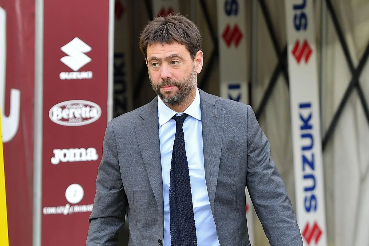 Uomo in abito grigio, camicia azzurra e cravatta scura davanti a banner sponsor Suzuki, Beretta e Joma