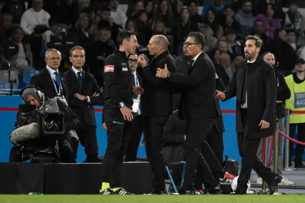Confronto acceso a bordo campo tra un uomo in abito scuro e un arbitro in tuta nera durante una partita di calcio