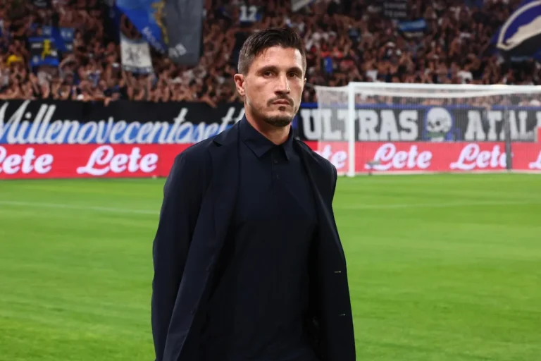 Uomo in abito scuro davanti a una curva piena di tifosi e a uno striscione Ultras, su un campo da calcio