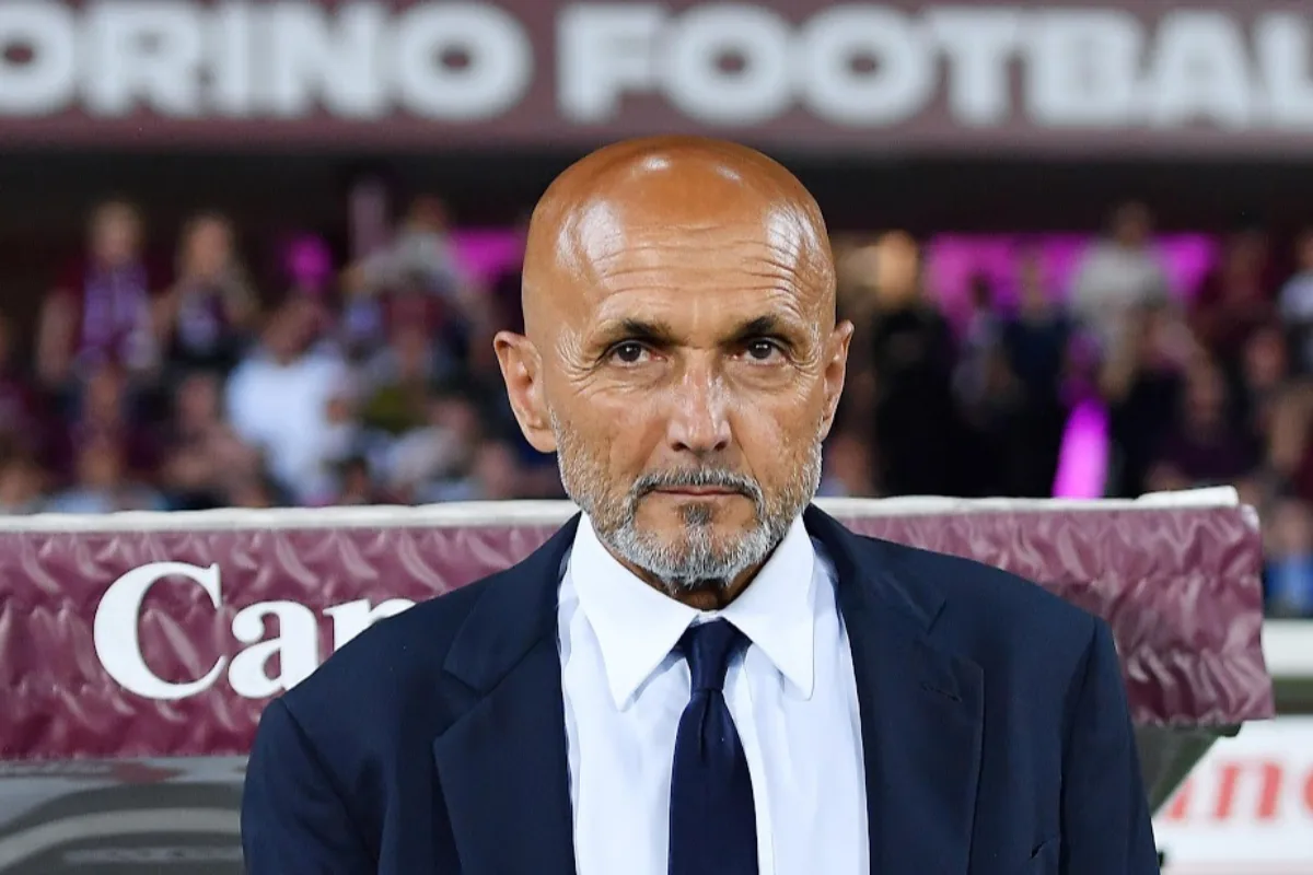 Spalletti con giacca blu e cravatta scura davanti a una panchina, con striscione Torino Football sullo sfondo