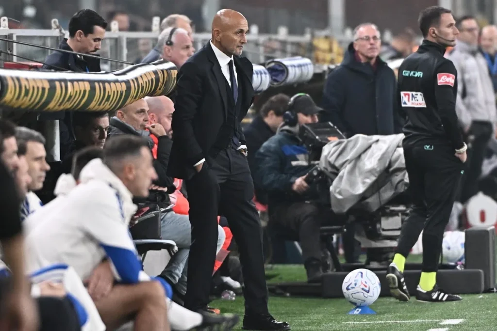 Spalletti in abito scuro a bordocampo durante una partita di calcio, accanto a un pallone sull'erba