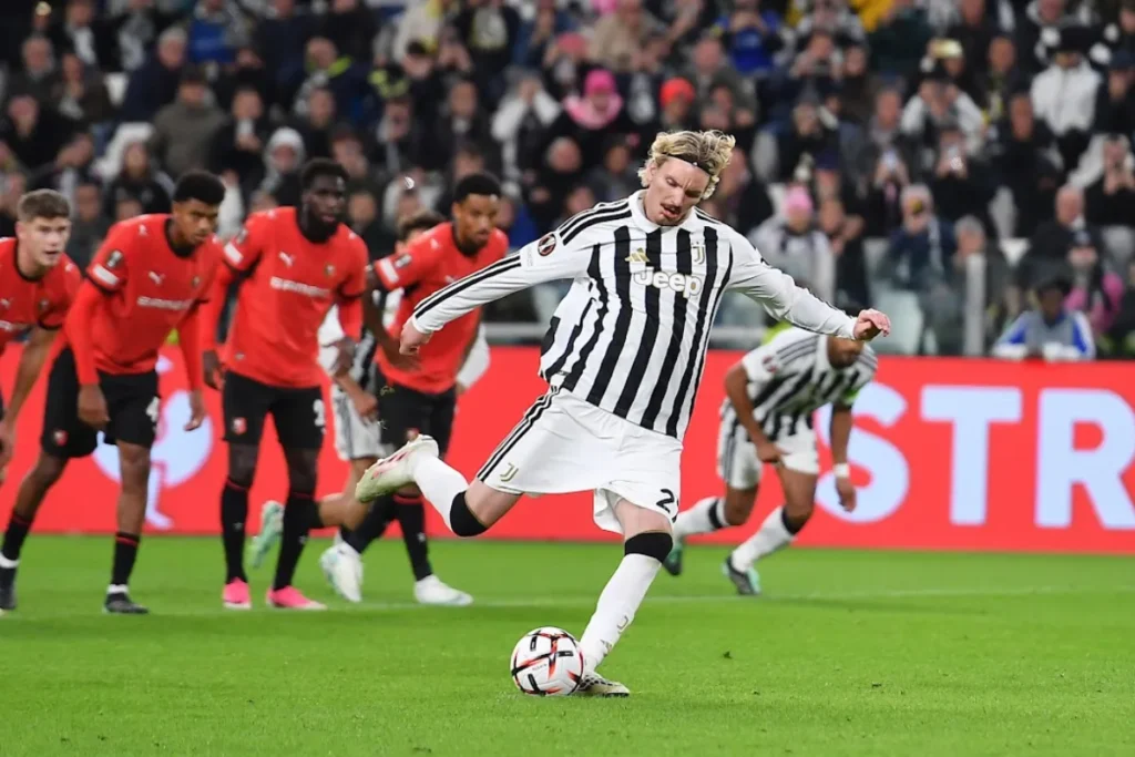 Giocatore della Juventus con maglia bianconera a righe calcia un rigore davanti agli avversari in maglia rossa, con il pubbli